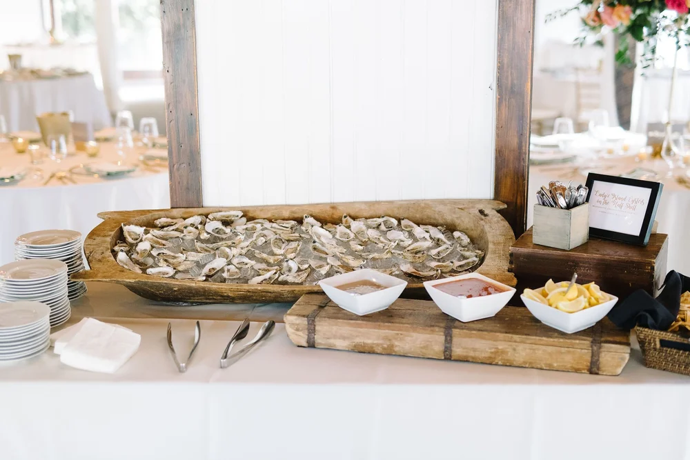 A buffet table with a wooden tray of raw oysters on ice, dipping sauces and lemon wedges at a Salthouse wedding